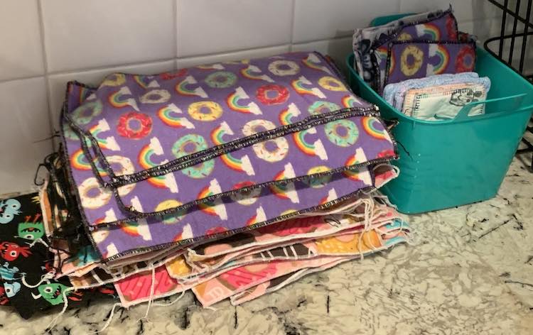 A stack of napkins sit on the counter against the backsplash with the cotton woven ones at the bottom. Beside it sits a teal plastic basket holding the smaller napkins and some other fabric items.