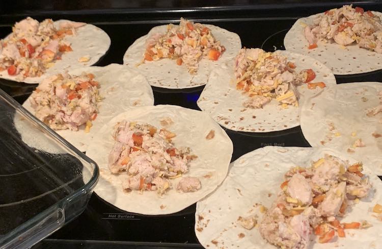 The black stovetop with an empty glass casserole dish off to the side, mostly off-screen, and white flour tortillas laid out over the rest. Each tortilla wrap 