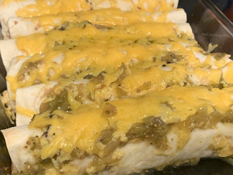 Closeup of the wrapped enchiladas lined up next to each other. They're covered in salsa verde, orange melted cheddar cheese, and the ends have browned a bit.