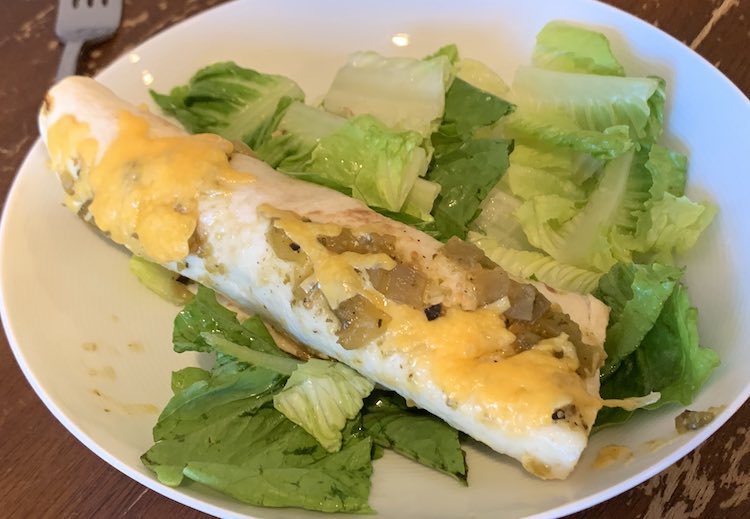 White bowl on a brown table with a fork off to the side. Inside the bowl is lettuce with a chicken enchilada placed overtop. 