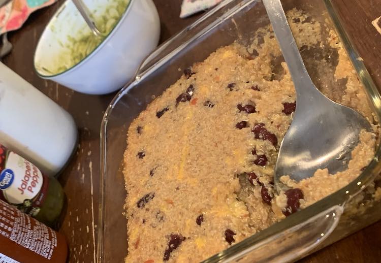 The square glass casserole dishes sitting on a hot pad on the kitchen table with a metal spoon in it after serving some of it out. Off to the side you can see the white bowl of guacamole, hot sauce, and pickled jalapeño peppers.