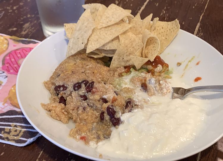 White bowl with the quinoa mix on the left, plain white yogurt on the right, guacamole/jalapeños/hot sauce in the back covered in tortilla chips. To the left is a fabric napkin with donuts on it and a glass of water. 