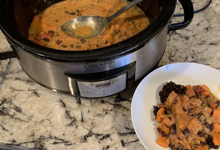 View of the finished stew in the crockpot and plated in the bowl. The bowl is in the lower right side on black rice while the crockpot sits in the upper left corner with cooked stew in it. 