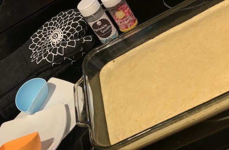 Image shows most of a long rectangular glass casserole dish a third filled with a smooth yellow pancake. Off to the side, back and left, are the waiting saucers, dip holder. oven mitts. and sprinkles.