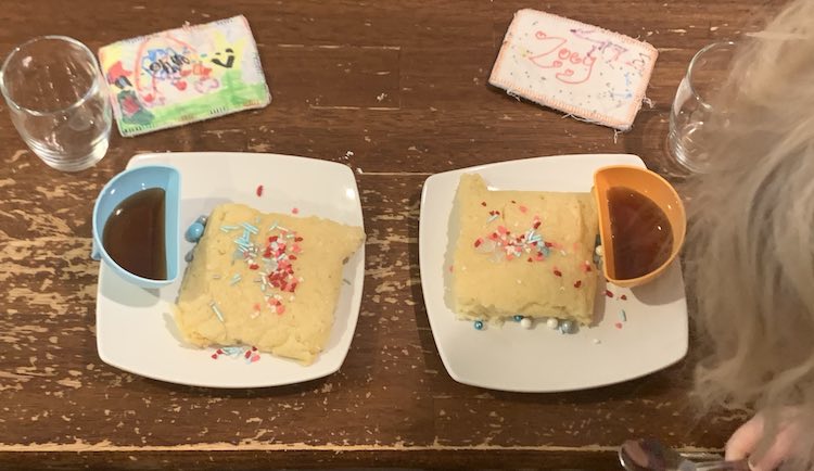 Overview of the two saucers of pancakes and syrup filled attached bowls. Zoey's head is starting to block one of them. Beside the plates are cups and their name tags.