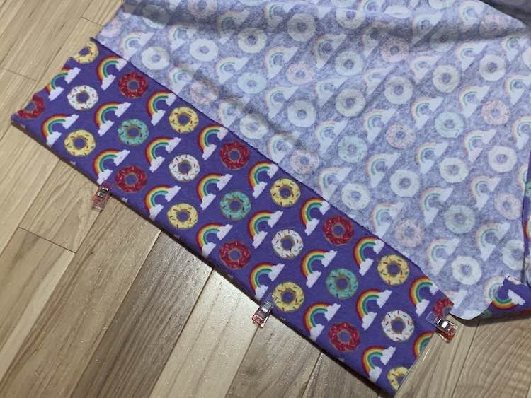Purple fabric with rainbows and donuts lay on the ground, wrong side up, with the end folded up and clips holding it in place.