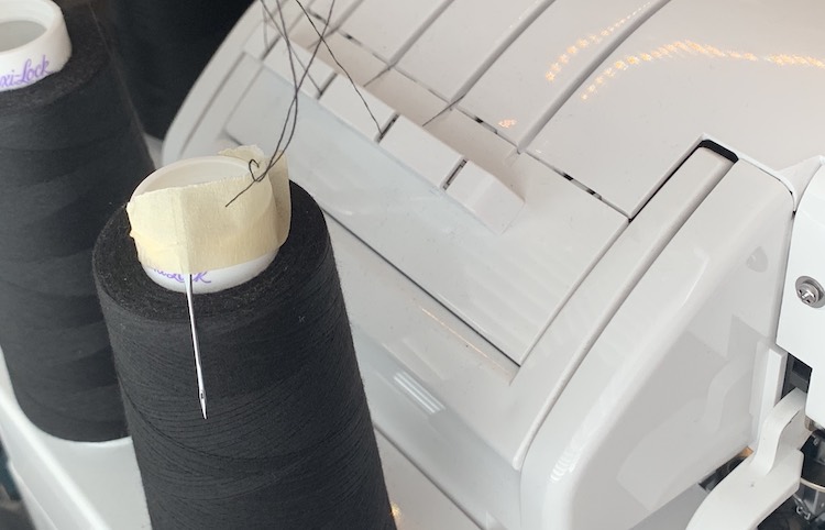 Closeup view of the back of my Babylock serger showing the one and a half cones of serger thread and the thread guides on the top of my machine. The closest spool of thread has a machine needle taped to the top of the cone with masking tape. 