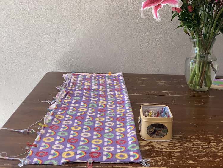 The photo shows the kitchen table with the purple fabric (with rainbows and donuts on it) folded in half. There's a container of sewing clips next to the fabric and some of the clips are attached along the left side and either end of the fabric. To the right top of the photo there lies a a vase with the bottom part of a lily shown. 