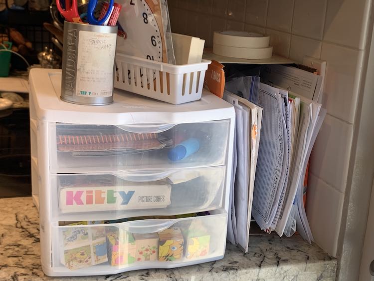 View of the counter with the stove behind it. Against the wall sits the paper organizer with two rolls of masking tape on it. Beside it sits the plastic drawers with a old David's Tea tin with dry erase markers and scissors alongside the white basket with a clock and other school items. In the drawers you can see the water wows and puzzles.