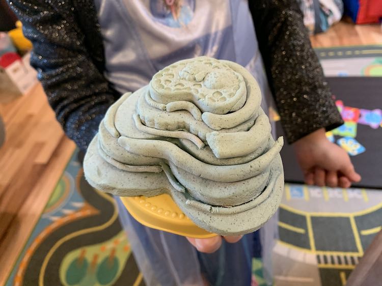 Image shows Ada holding up a plastic cake stand with layers and layers of hardened blue hearts forming a playdough cake with a strawberry shape on top. Behind her the living room is slightly blurred. 