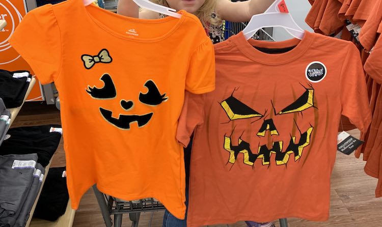 Image shows Zoey sitting in a shopping car at Walmart. She's holding up two shirts that, mostly, covers her up. The one on the left is a brighter orange, thinner fabric, with a cuter jack-o-lantern shapes and bow. The one on the right is slightly scarier with pumpkin curved vertical lines and a sticker letting you know it glows. 