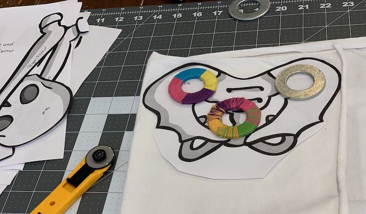 Image shows the cutting mat with a section of folded over fabric with the pelvic bone placed overtop with three washers holding it down (two wrapped in embroidery floss). Off to the side there's another washer, a rotary blade, and a pile of paper bones. 