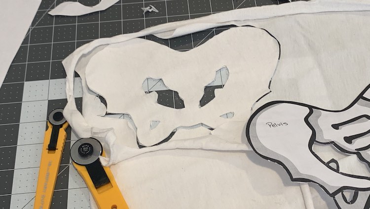 Image shows the cut out fabric pelvis with the empty spaces emptied and the fabric around it slightly pulled away. The cutting mat is visible under the fabric while the paper pelvis and the two rotary cutters sit below. 