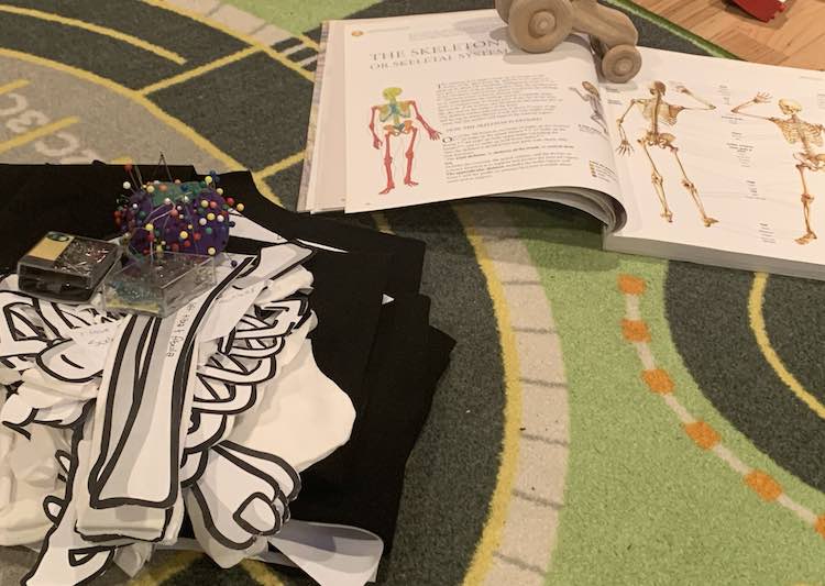 Laid out on the car mat sits an opened book to the skeleton section with a wooden vehicle laid overtop to keep it open. The bottom left of the image shows a pile of black fabric, with the odd visible sliver of white paper, with a pile of the white bones overtop. Perched on top is a purple tomato with pins in it and two extra containers of pins just in case it's needed. 