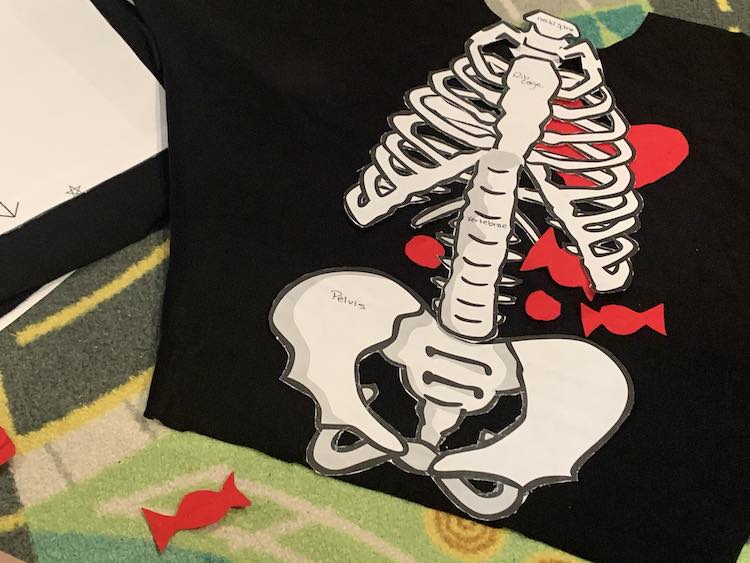 Image shows the shirt opened up over a car mat. On the shirt sits the paper bones for the neck, rib cages, spine, and pelvis. Scattered around under the bones are the candy and heart. 