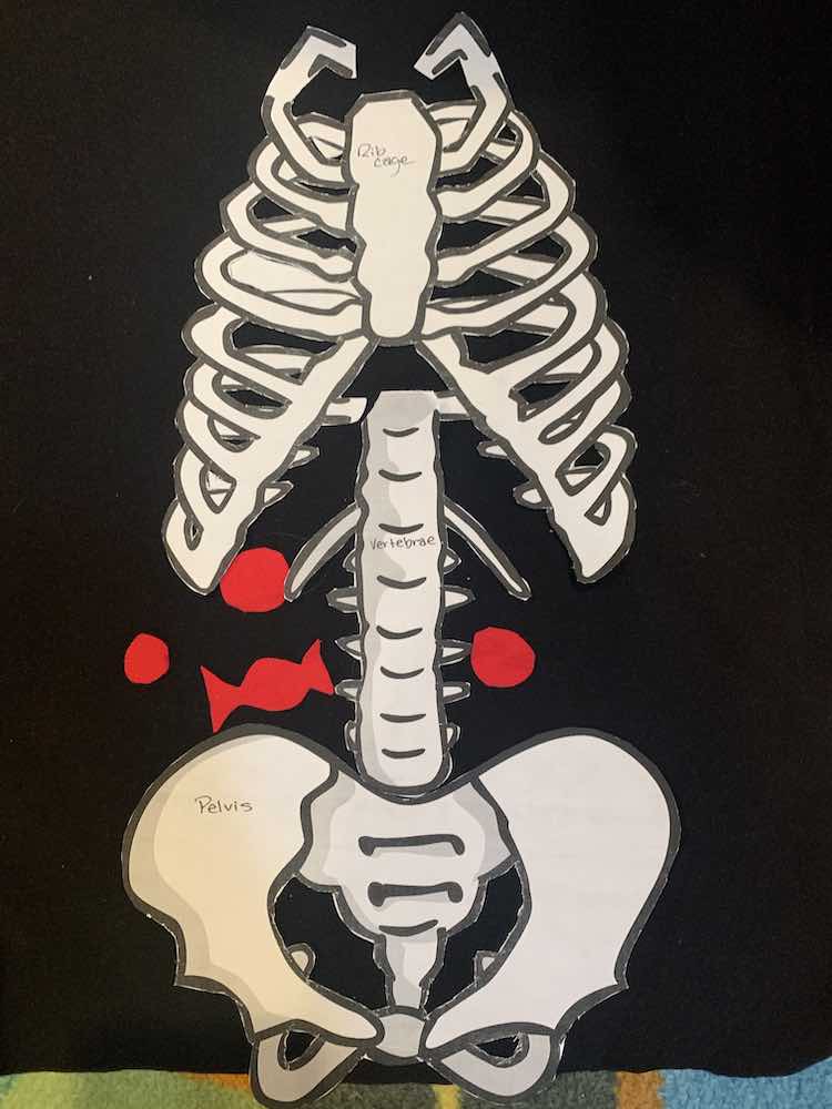 Image shows a closeup of the black shirt with the paper bone pieces laid overtop. There are four red candies placed around the vertebrae.