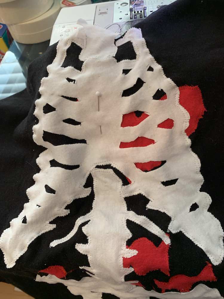 Image shows a closeup of the front of the shirt close to the sewing machine. You can see the stitching around the outside of the neck, ribs, spine, and pelvic bone. There's still a pin in the center of the ribs but all the other pins were taken out.