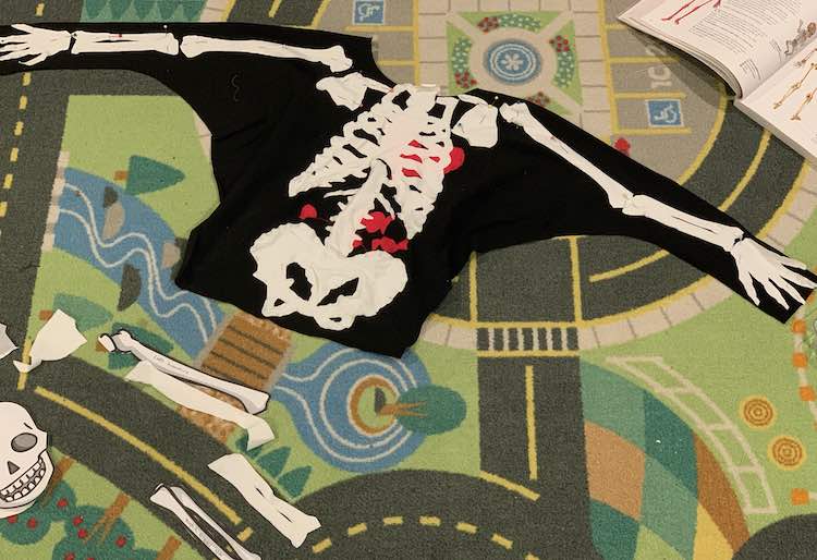 Image shows the car mat with the front of the shirt laid out on it. All the center bones, candy, and heart are sewn on while the arm, and related, bones are pinned on. Below you can see the extra bone pieces that haven't been used and above it the opened body book with the bottom of the skeleton diagram showing.