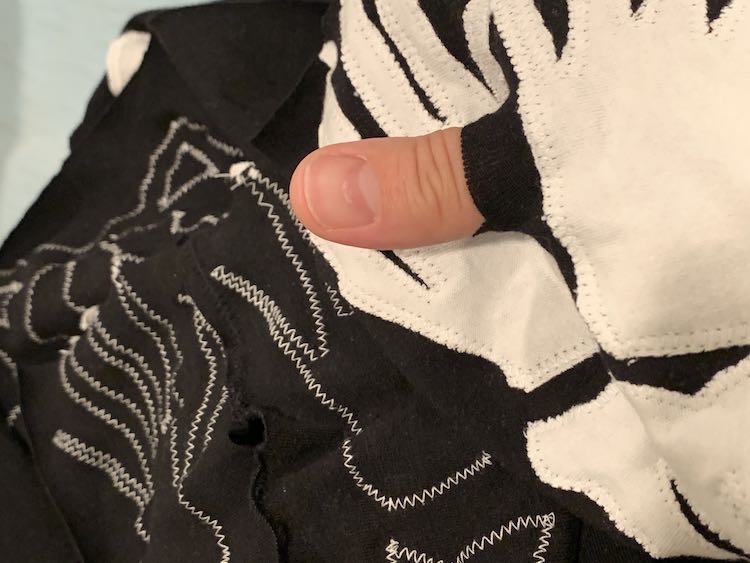 Image shows multiple hands sewn onto the shirt. On the right, on the foreground, are two white hands sewn onto a black background. My thumb is sticking out showing the edge of the fabric to show how the thumbhole works. In the background, to the left, you can see the underside of the sleeve with the thumbhole pointed inside. Since it's the underside you can't see the white fabric but can see the white thread zigzagging the outline of the hands on the reversed side. 