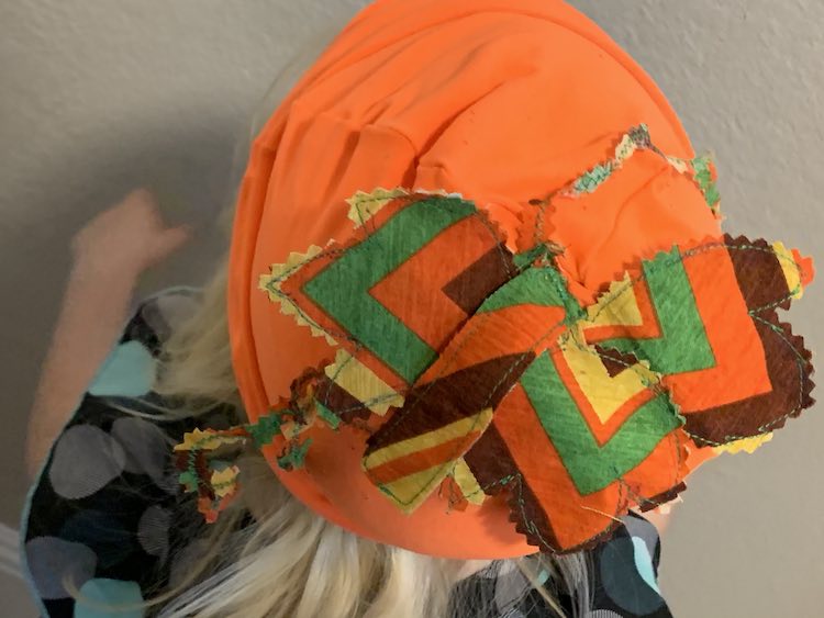 image shows the top of the beanie with the stem, leaves, and vines, mostly, drooping down. In the background is a white wall and the top of her body slightly blurred. 