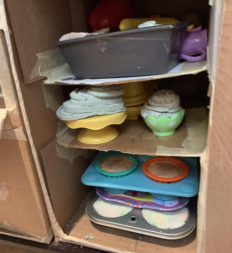 Image shows cardboard shelves surrounded by more cardboard in a table. The shelves are all filled with drying out playdough placed in either muffin tins, cake platter, loaf pan, or half a plastic Easter egg.  