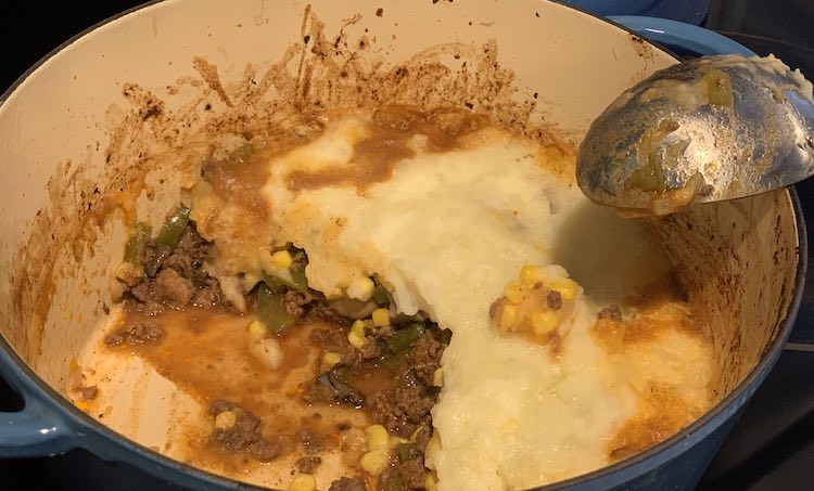 Photo taken from above looking down at a half filled dutch oven with shepard's pie. Along the edge you can see the ground beef mixture, with a dot of yellow and green, spilling out from the white topped section. A serving spoon sits leaned up against the pot edge.