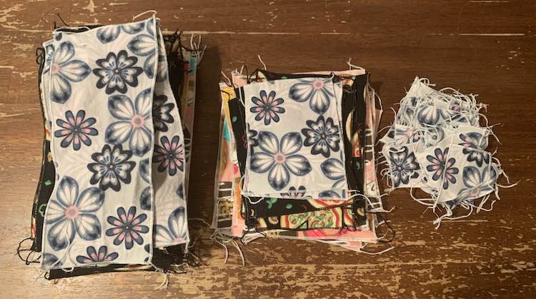 There are three piles on the table and the photo is take from above looking down on the piles. Each is topped with the grey flowered fabric though you can see the other two fabrics peaking out on the two left piles. The leftmost pile consists of large rectangular napkins, the center pile are smaller square napkins, and the rightmost pile are small single-layered face cloths. 