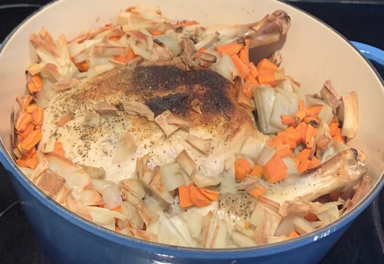 Angled overview of the inside of the dutch oven containing a whole chicken. The browned and seasoned top of the chicken and tips of the drumsticks are sticking out from it's diced white potato and orange carrot bath. 