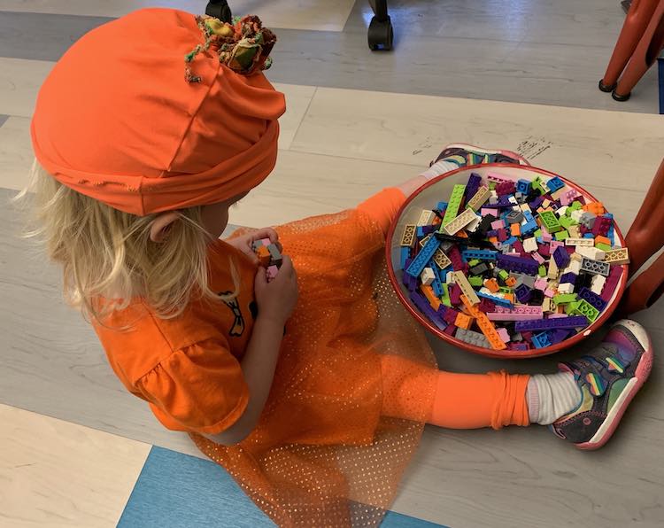 Photo taken a bit above and to the side of Zoey. She's sitting on the floor focused on putting some LEGO bricks together. Between her feet sits a bowl full of more LEGO bricks. 