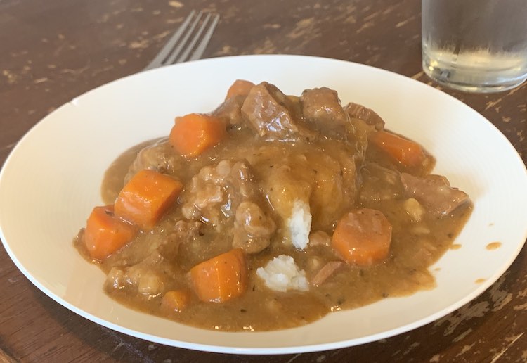 White plate with a fork and glass of water off to the side. On the plate is a pile of white potatoes covered in brown-ish liquid and chunks of meat and carrots. 