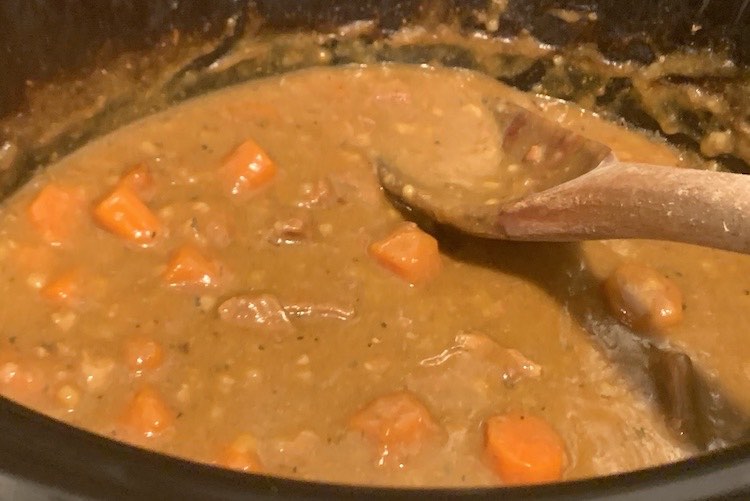 Closeup looking into the slow cooker with a wooden spoon leaned inside. The contents are orange browny with floating chunks of beef and carrot inside.