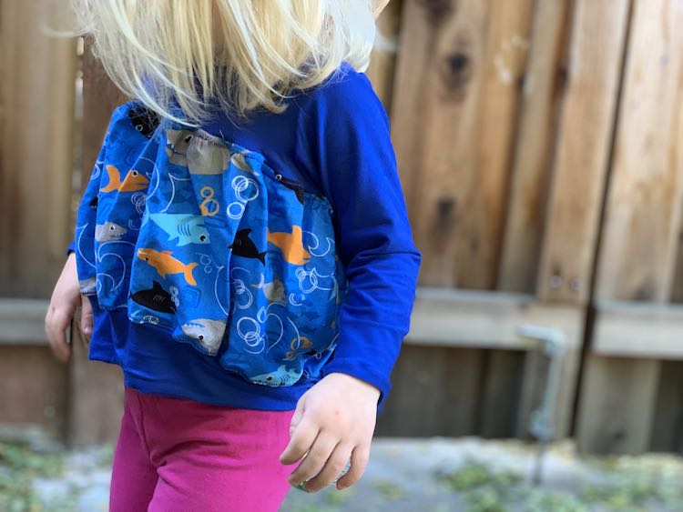 Image shows a closeup of a blue long sleeved top with a puffed up shark fabric over the midsection. It's paired with pink pants with the background fence blurred.