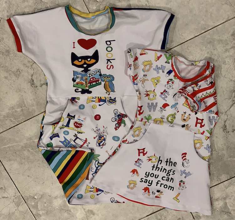 Flat lay of both dresses laying on the kitchen floor. One corner of either dress is flipped over to see the back fabric. The larger dress has Pete the Cat holding a book beside an owl saying "I <3 books". The back and the bands are striped, around the image is white, and the bottom of the skirt shows scattered images from Pete the Cat. The smaller dress, in front, has the panel on the skirt saying "Oh the things you can say from A to Z". The rest of the fromt of the dress has Dr Suess-eque letters whiel the black is white with red strips and characters on the white. 