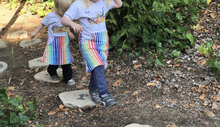 Front view of the matching dresses as the girls walk over mathematical stepping stones (they count from one to five in different small equations). Ada is in the front and you can see from the top of her dress down. Behind her and partially overlapped is Zoey in the slightly more oversized dress. 