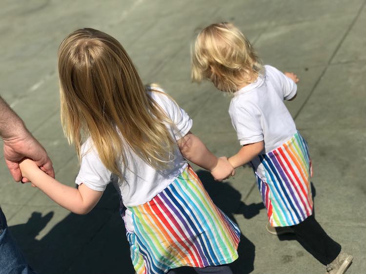 View from the back of the dresses as the girls hold hands. You can see Matt's hand to the left holding Ada's hand. On the other side Ada is holding Zoey's wrist as they walk. 