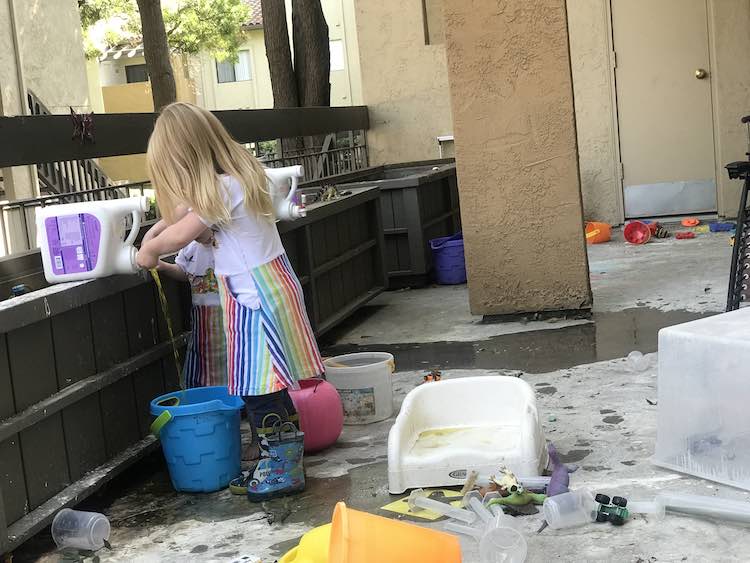 View of a mess covered deck at our old apartment. The kids are gathered around an old laundry detergent bottle balanced on our garden ledge. You can see part of Zoey's dress and the side of Ada's as Ada presses down on the bottle button so dyed yellow water dribbles out into a bucket. There's old plastic test tubes on the ground so this happened sometime after her science party.