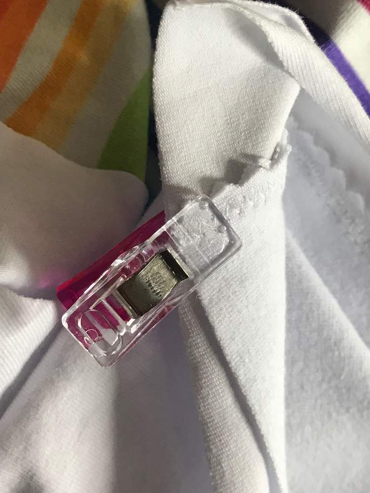 Image shows a closeup of one end of the zigzag trimmed seam. At the end of the seam there's a clear and pink sewing clip holding the seam flat against the fabric. In the background is the rest of the white knit garment slightly blurred in places. 