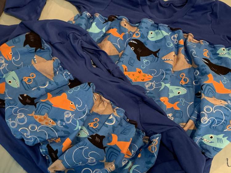 Image shows two shirts laid partially on top of each other on the ironing mat. Both garments have dark blue knit at the top and bottom with blue shark filled fabric in the center. The smaller shirt has one grey shark with orange hearts drawn on it while the back shirt has an orange shark with two hues of blue hearts on it. 