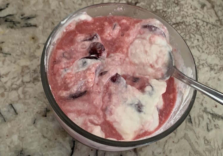 Closeup image showing a circular glass container filled with chunks of red, white yogurt, and pink 'yogurt' where it mixed. A metal spoons is poking out of the yogurt.