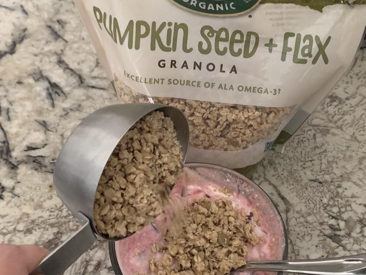 Image shows a glass circular container with mixed cherry and yogurt inside it. The yogurt is at the bottom of the photo with a bag of Pumpkin Seed + Flax Granola sitting behind it. There's a metal measuring cup with granola over top of the yogurt and it's tilted so the granola is pouring onto the yogurt. 
