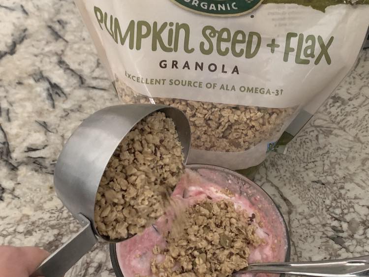 Image shows a glass circular container with mixed cherry and yogurt inside it. The yogurt is at the bottom of the photo with a bag of Pumpkin Seed + Flax Granola sitting behind it. There's a metal measuring cup with granola over top of the yogurt and it's tilted so the granola is pouring onto the yogurt.
