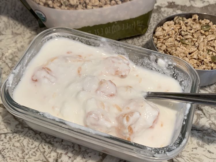 Photo shows a glass rectangular container filled with white yogurt and pink chunks of grapefruit. There's a metal spoon sticking out of the container. Behind the yogurt is a bag of granola, to the left, and, to the right, a metal measuring cup filled with granola.