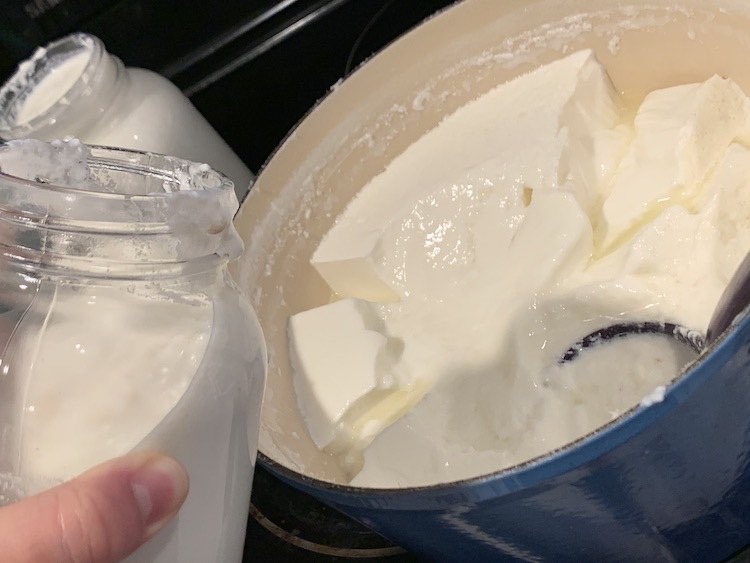 Shows my hand holding and tilting a 32 oz. canning jar over Dutch oven. There's a filled canning jar in the background. Inside the Dutch oven are clumps of yogurt within the whey liquid and you can barely see the purple plastic soup ladle sitting among the yogurt. 