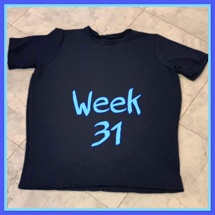 Image shows a simple black t-shirt laid out on the tiled floor with the words "week 31" in blue over the center. The image is outlined in light and then dark blue.