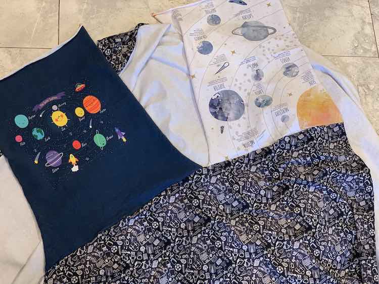 Image shows the coordinating black doodle fabric, along the bottom, with the two panels connected at the top. The panel on the left, navy blue with planets, if right side up while the panel on the right, white with a yellow sun surrounded by planets and descriptions, is upside down. The panels are only connected on the one side and not on any other side.  