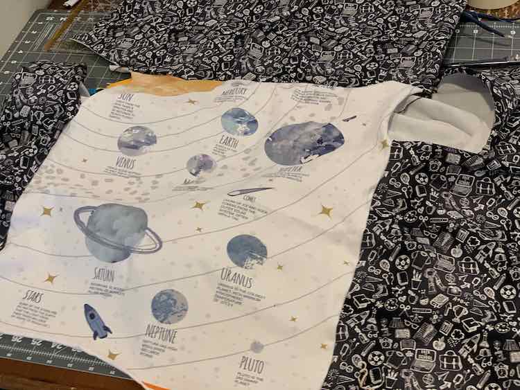 Image shows the white panel with blue planets with the bottom curling up and the top, and most of either side, extended by the black fabric with white school-related doodles. Underneath it all is a cutting board. 