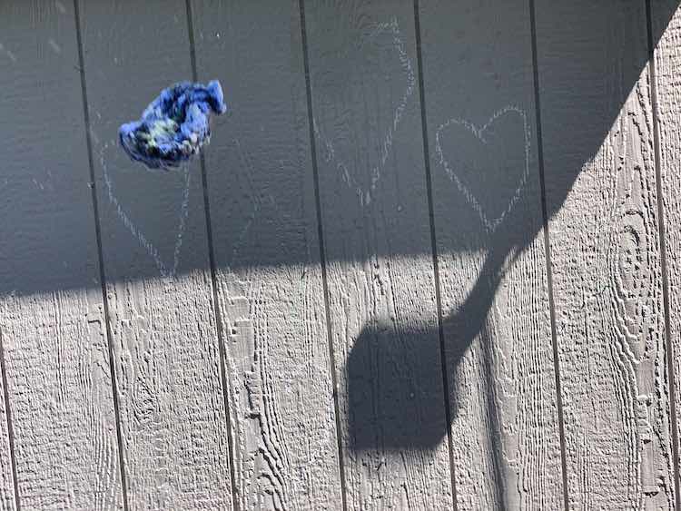 Image shows a shadow covered shed with some white outlined shapes drawn onto the wall. In the foreground is a single wet yarn water balloon flying towards the wall and spraying water out the back (above). 