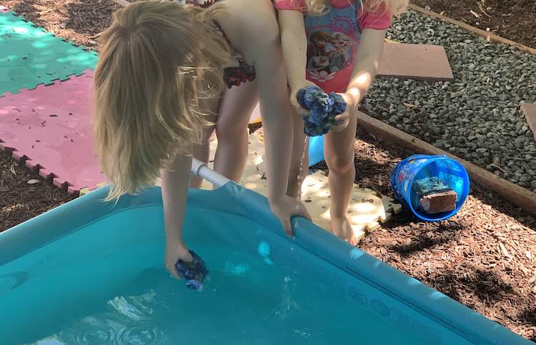 Both kids are standing behind the small pool. Ada is slightly in front and has one hand on the side of the pool as she bends over to soak the water balloon in her other hand. Zoey is slightly behind her and is squeezing her balloon out over the pool. 