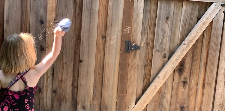 Background of the image shows a wooden fence with a couple small orange circles colored on. Ada stands in front of it with a single arm raised holding a wet water balloon. You can see she started swinging as there's water flying off of it towards her in an arc. 