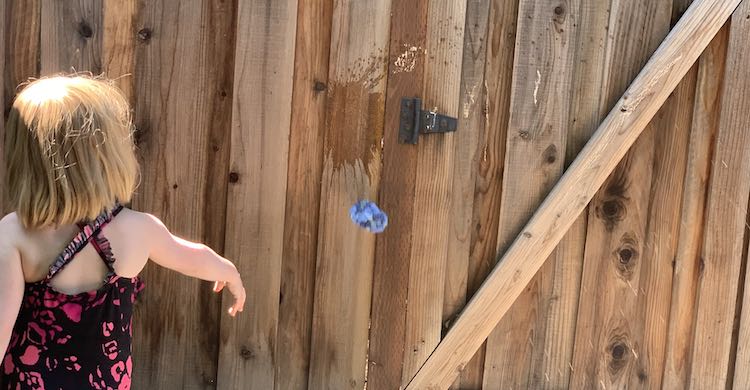 Ada's arm is now outstretched and angled downwards as she just finished throwing the ballon. The balloon is crumpled up a bit and is falling to the ground. Slightly above the balloon, on the fence, there's a wet splat mark where one of the sidewalk chalk targets were.